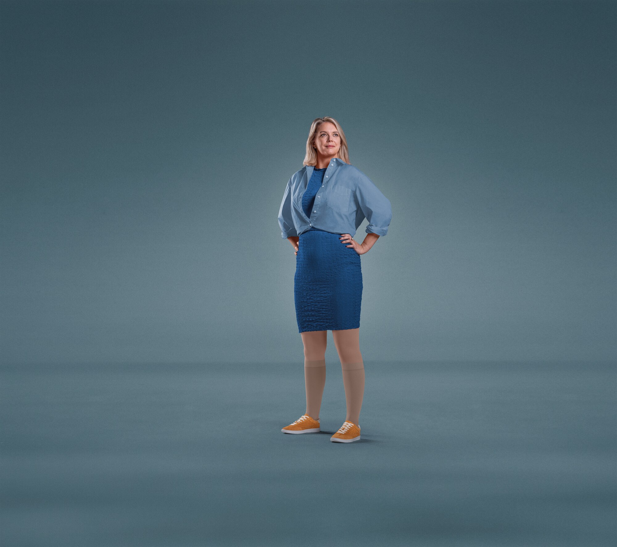 A woman standing against a plain blue background with both hands on her hips, dressed in a blue shirt, blue skirt, and orange shoes, wearing Knee-high JOBST Relief compression stockings.